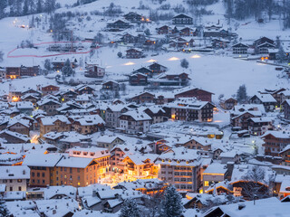Snow-covered Moena village, Fassa Valley, Trentino, Italy. Lights glow from windows and street lamps, illuminating the winter scene at dusk.