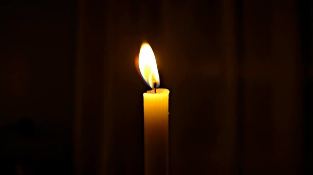Illuminated paraffin wax candle with a visible burning flame against a dark background providing light with soft ambient glow.