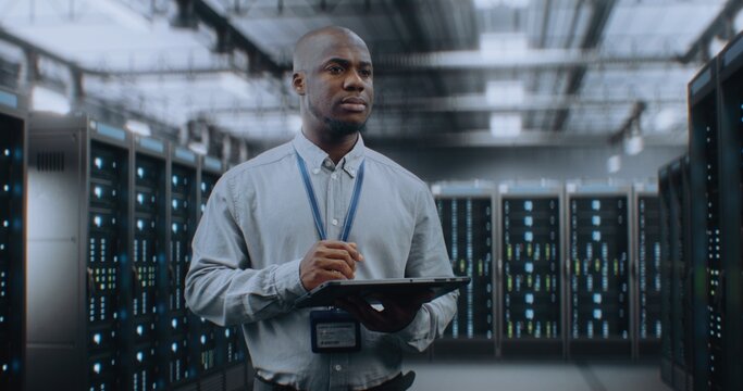 Big Data Center with Server Racks: Professional African American Specialist Using Tablet Computer, Activating Servers, Standing In Warehouse. Information Technology and Cyber Security. Medium Shot.