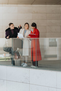 Three women on an office balcony reviewing documents while engaged in a discussion, symbolizing female empowerment and professional collaboration in a corporate environment.