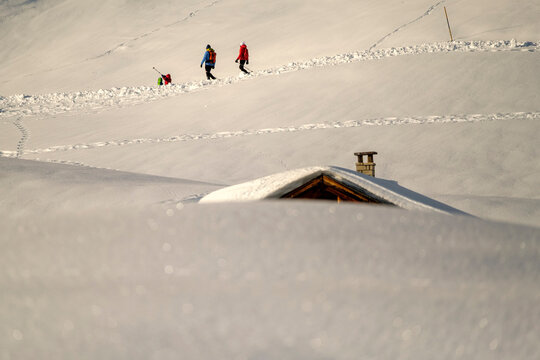 Hikers trek through deep snow past a partially buried cabin. Fuciade, Fiamme Valley, Trentino, Italy. The roof and chimney are visible above the snow.