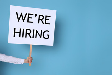 Man holding sign with phrase We Are Hiring on light blue background, closeup. Recruitment, human resources concept