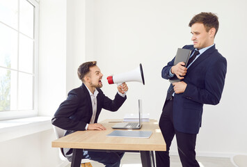 Plakat Angry boss sitting at the desk on his workplace holding mouthpiece and shouting at his young stressed employee standing in office. Businessman yelling at male worker. Dismissal concept.