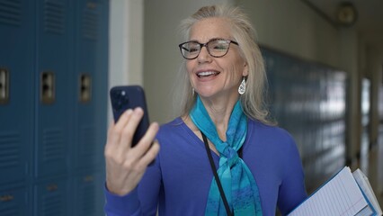 A mature woman, dressed professionally, happily having video chat phone in a school corridor filled with lockers. She holds a notebook and a pen.