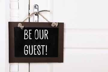 Small blackboard with phrase Be Our Guest hanging on door handle, space for text