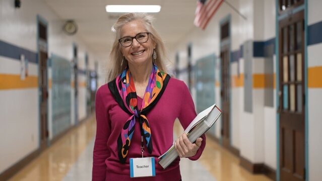 A mature woman teacher smiles as she walks confidently through a school hallway. Holding a stack of files she wears a colorful scarf and has a name tag visible ready for her class.