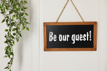 Small blackboard with phrase Be Our Guest and houseplant hanging on white wall