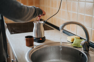 Person stirring a hot drink in a metallic kettle on a countertop, with a clean ceramic kitchen sink and warm tiled back splash. A relaxed domestic setting with an emphasis on comfort.