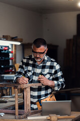 Carpenter standing in his workshop restoring old piece of furniture marking measurements on wood...