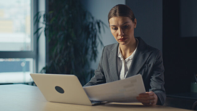 Business executive or manager in office reading a document with serious expression, sitting at desk with laptop. Concept of responsibility, analysis, decision-making, focus, and professional duty - Powered by Adobe
