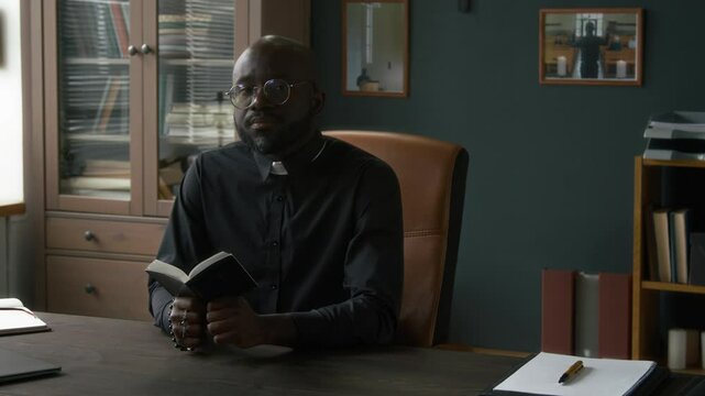 Portrait of bearded Black priest dressed in clerical shirt posing for camera while reading small bible book at work table in church office