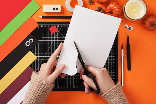 Seasonal diy paper crafting. Hands cut a ghost silhouette out of white paper with scissors. Around paper crafting items, pencil, tape, cutter, tweezers. - Powered by Adobe