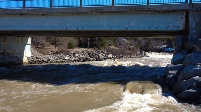 A river with brown water resulting from the spring flood flows under a road bridge made of metal girders and cement pillars. Tartigou River, Quebec, Canada, 2025.