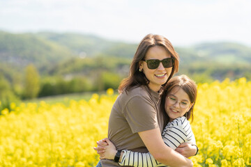 Daughter and mother hugging and smiling standing in front of blooming yellow rapeseed field....