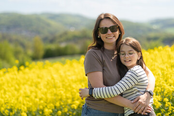 Daughter and mother hugging and smiling standing in front of blooming yellow rapeseed field....