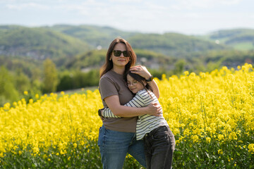 Daughter and mother hugging and smiling standing in front of blooming yellow rapeseed field. Concept of family emotions and leisure