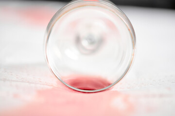 Empty wine glass lies on white tablecloth stained with red wine. Soft lighting creates gentle shadows, highlighting contrast between glass and fabric. Close-up angle emphasizes detail
