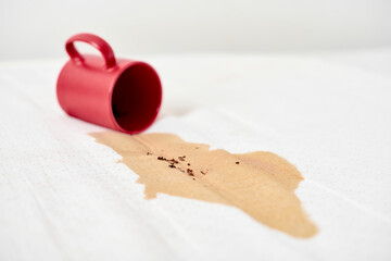Red mug tips over, spilling coffee on white surface. Brown liquid stain spreads, creating contrast...