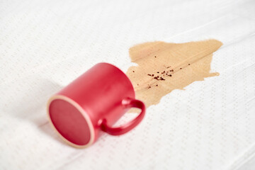 Red mug lies on white fabric, brown liquid spilling out. Warm lighting casts soft shadows. Coffee...