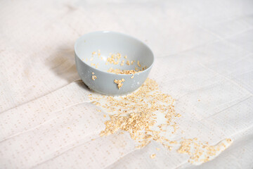 Light blue bowl with remnants of oatmeal tipped over, spilling contents onto white fabric. Soft...