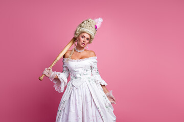 Graceful woman in a baroque style dress holding a cricket bat against a pink backdrop representing a blend of modernity and historic charm
