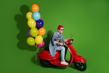 Stylish woman on red scooter holding colorful balloons against vibrant green background, enjoying...