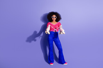 Stylish young woman in vibrant retro fashion holding roller skates against a purple background expressing joy