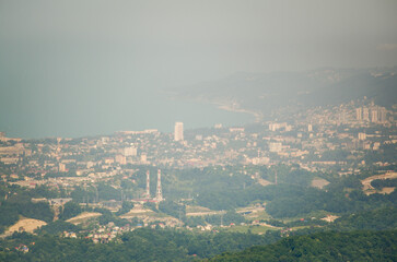 Cityscape viewed from high angle, surrounded by verdant hills. Soft daylight creates gentle contrast between urban structures and natural greenery. Background hazy with distant urban skyline