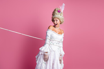 Sophisticated woman tied with rope dressed in royal baroque costume humorously posing on pink...