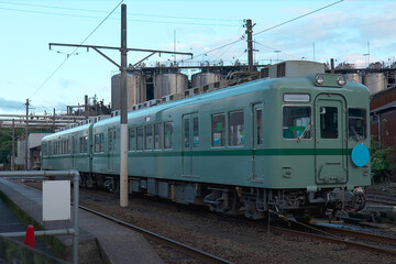 Fototapeta premium Vintage green Japanese train parked beside an industrial complex, with tanks and pipes in the background under a clear blue sky. 