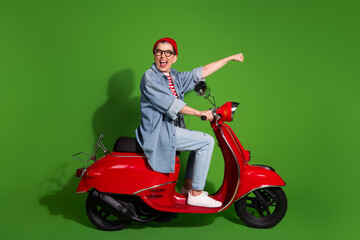 Stylish senior woman in a casual fashion outfit riding a red scooter against a vibrant green...