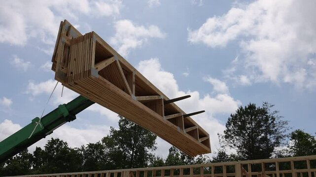 Forklift telehandler machinery raises wooden trusses beams high in air as fluffy clouds fill blue sky during workday.