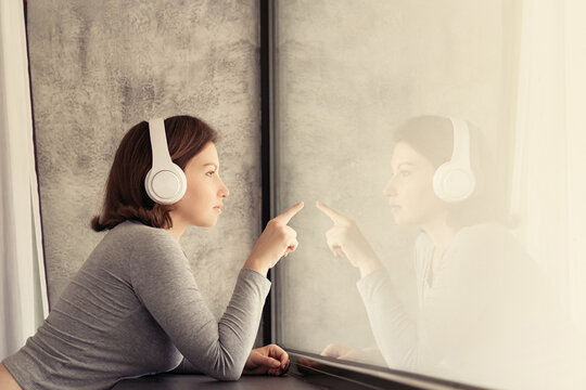 Portrait of beautiful сaucasian young woman in headphones touches her reflection of big panoramic window of apartments and listening music.