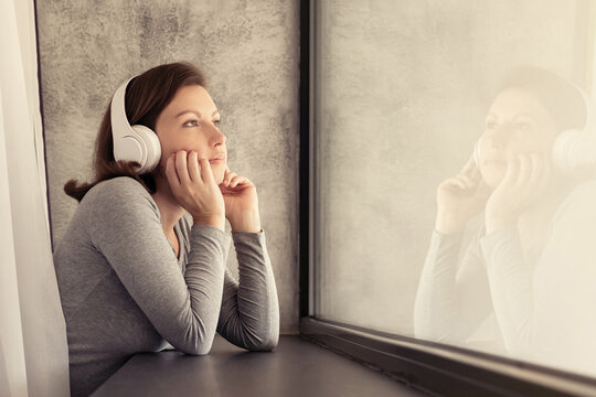 Portrait of beautiful сaucasian young woman in headphones dreaming, enjoying listening music standing and looking near big panoramic window of apartments.