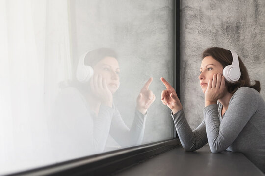 Portrait of beautiful сaucasian young woman in headphones touches her reflection of big panoramic window of apartments and listening music.
