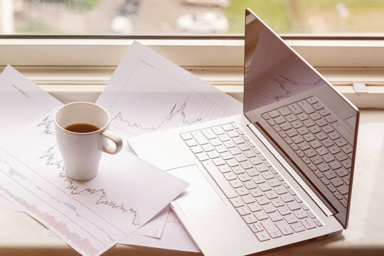 cup coffee,laptop with graphics and charts printed on the paper lying on the windowsill near the window of apartment. remote work concept