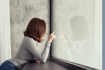 Portrait of beautiful сaucasian young woman touches her reflection of big panoramic window of apartments and listening music.