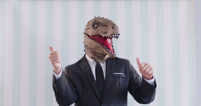 Creative man in brown dinosaur surreal rubber head mask and black suit showing thumbs up and dancing with his index fingers up. Weird guy having fun in the studio isolated on striated background.