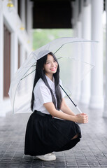 beautiful Thai university student woman in uniform squatting under a transparent umbrella during rainy season
