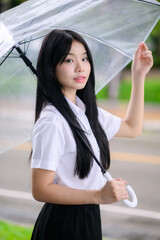beautiful Thai university student woman in uniform squatting under a transparent umbrella during rainy season