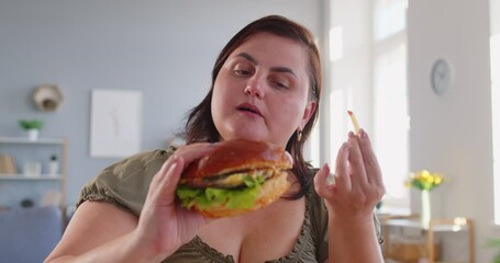 Close up of overweight fat woman enjoying big burger and french fries at home. Plus size female indulging in fast food, showing carefree attitude, love for tasty meal and unhealthy eating habits.