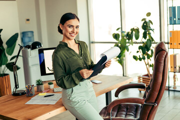 Confident businesswoman smiling in modern office setup with desk and stylish decor, holding...