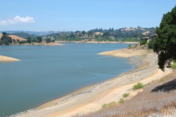 A reservoir with low water levels, exposed shoreline, and surrounding hills under a clear sky.