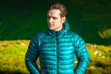 Portrait of a young man in a green down jacket looking away with serious face in windy mountain landscape © XArt