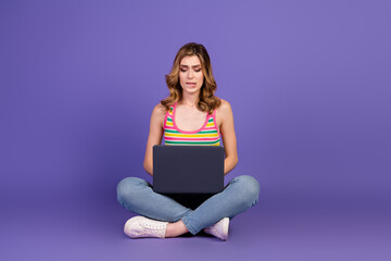 Young woman sitting with a laptop on a purple background, wearing casual clothes and a striped top