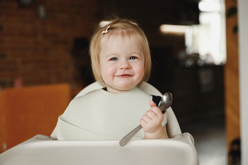 Infant girl or toddler sitting on high chair. Eating food by herself using spoon