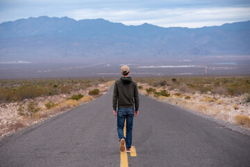 Obraz premium Man in a green sweatshirt and jeans walks alone on desert road toward mountains on Route 66 in Arizona.