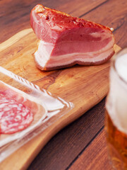 A slice of ham is on a wooden cutting board next to a glass of beer. The ham is cut into a triangle shape, and the beer is in a glass. The image conveys a casual and relaxed atmosphere, as the food