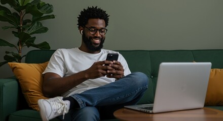 man using smartphone on green sofa with laptop nearby. Remote work and digital communication concept. Home office and online connectivity. Modern lifestyle and technology integration