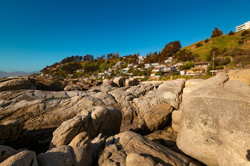 The scenic beach resort town of Maitencillo in Chile's V Region stretches across a panoramic view, beautifully contrasted by a prominent rocky area in the foreground, with the beach houses in the back
